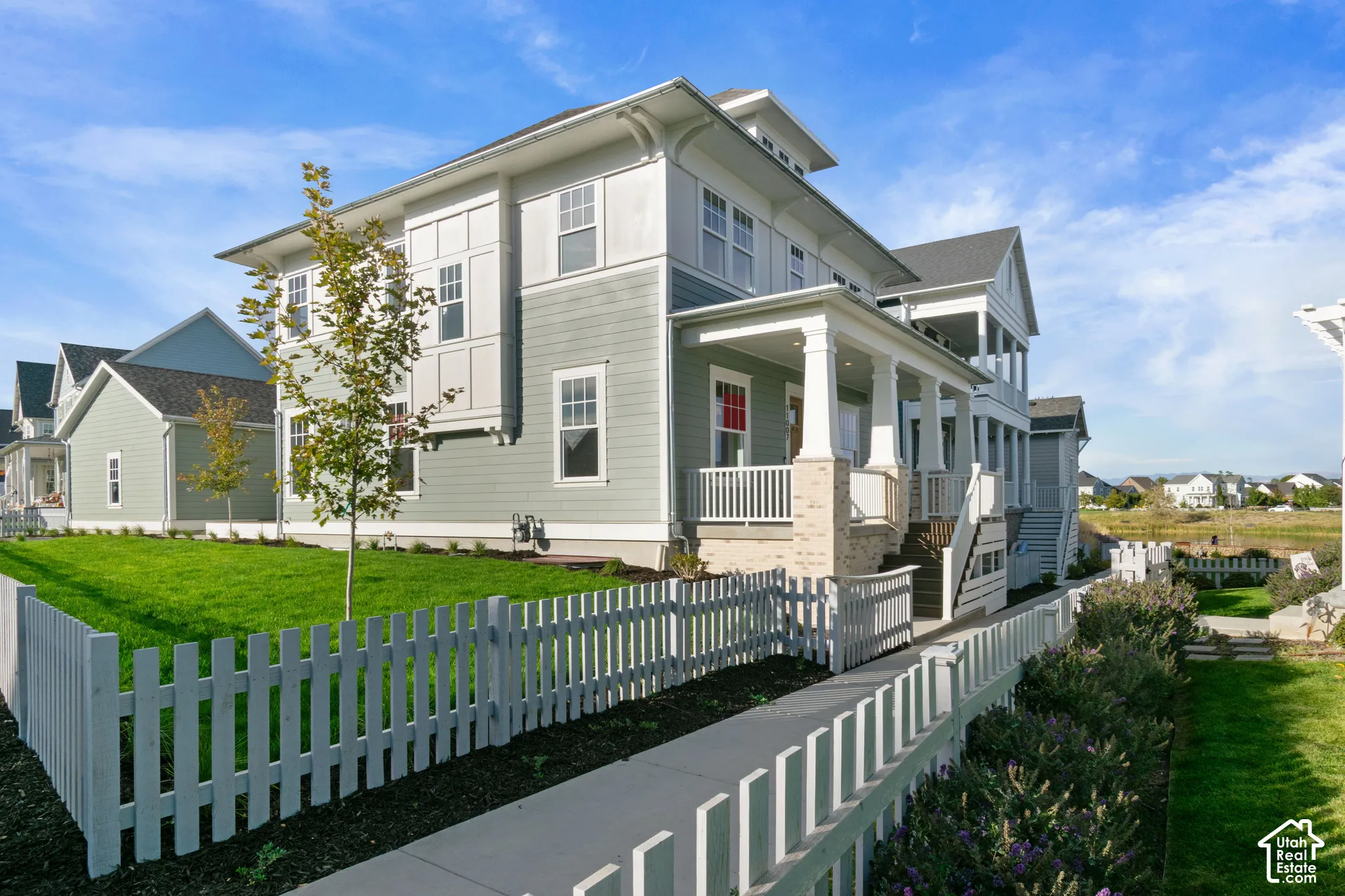 Multi-level exterior featuring light green horizontal siding, vertical board-and-batten accents, stone-base columns on the covered porch, and a white picket fence enclosing the manicured lawn