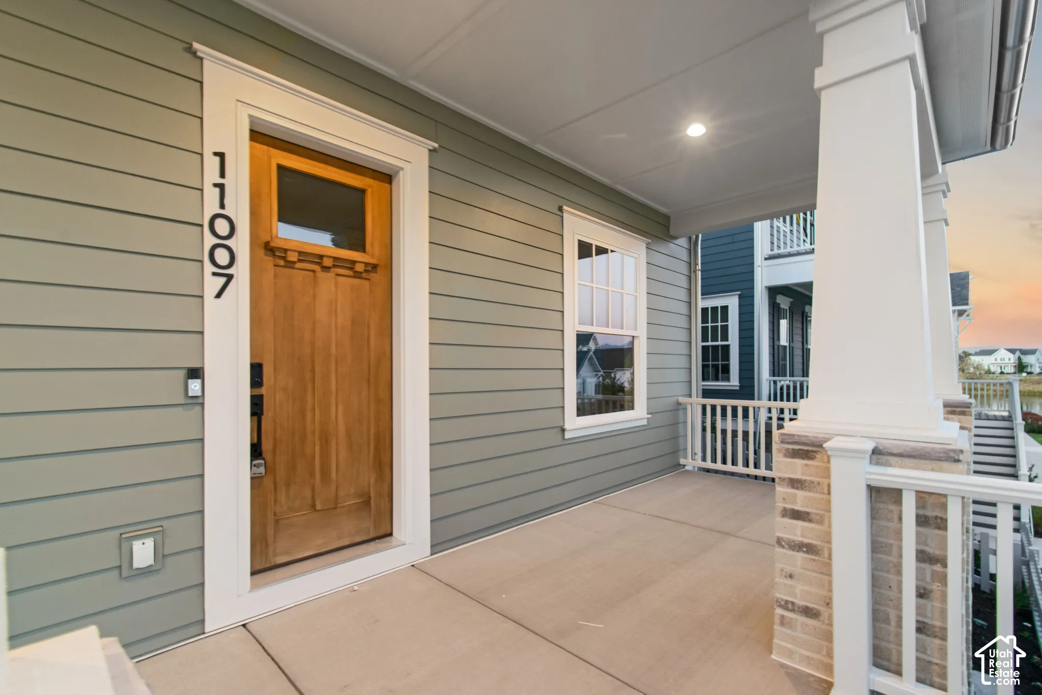 Covered front porch featuring horizontal lap siding, a wooden front door with an inset window, square structural columns, and a brick-based railing