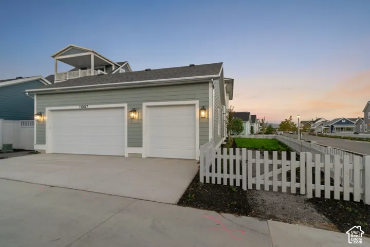 Two-car garage exterior featuring horizontal siding, white trim, wall-mounted coach lighting, and an attached white picket fence enclosing a lawn area
