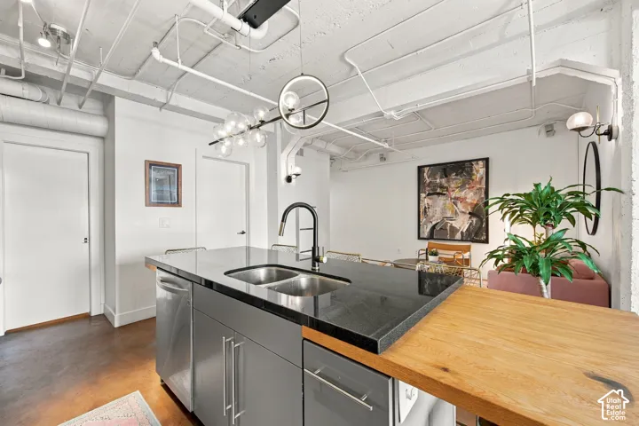 Kitchen island with a dark stone countertop, stainless steel double basin sink, integrated dishwasher, and a wood-finish breakfast bar