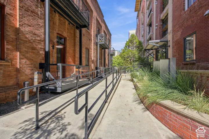 Concrete ramp with metal handrails, flanked by brick buildings and a planter with established grasses