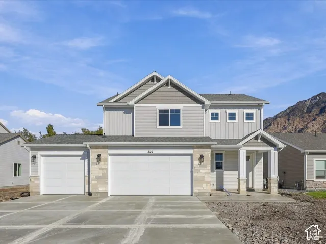 Two-story residence featuring light gray vertical and horizontal siding, stone veneer accents, a three-car garage, covered entryway with columns, and mountain scenery