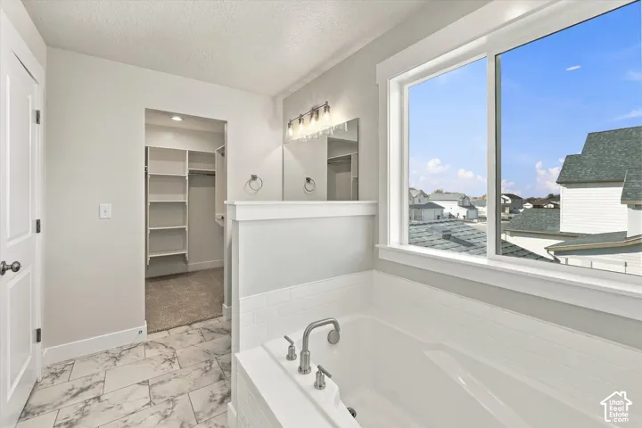 Soaking tub with a subway tile surround, white marble-finish flooring, multi-light wall-mounted vanity fixture, and walk-in closet with built-in shelving