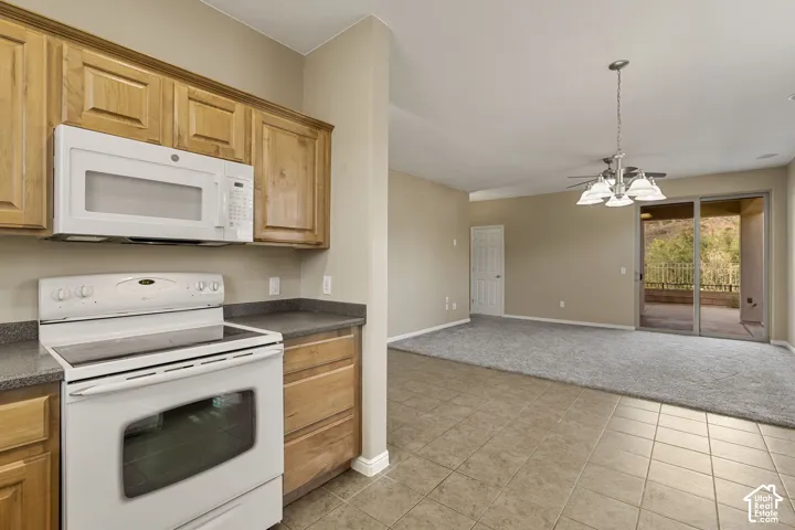 Kitchen featuring stained wood cabinetry, stone-finish countertops, and white appliances including a built-in microwave and range, alongside a tiled floor, five-light chandelier, and glass sliding doors