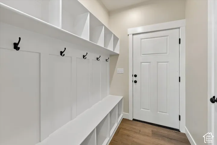 Built-in storage bench featuring partitioned cubbies, white wall paneling, five mounted matte black coat hooks, and overhead open shelving, accented by wood-finish flooring