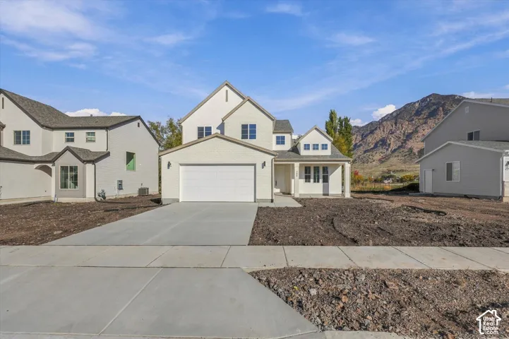 Two-story residential exterior featuring a light-toned brick facade, gabled rooflines, two-car garage, and concrete driveway, set against a mountain backdrop