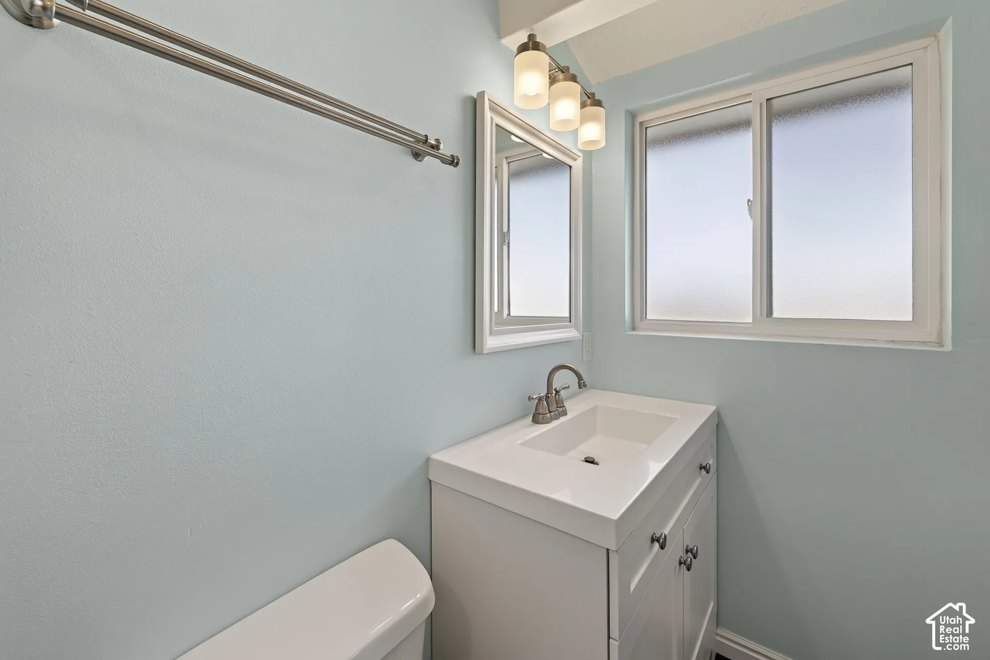 White vanity with integrated rectangular sink and metallic hardware, framed wall mirror, three-light vanity fixture, and horizontal window with frosted glass