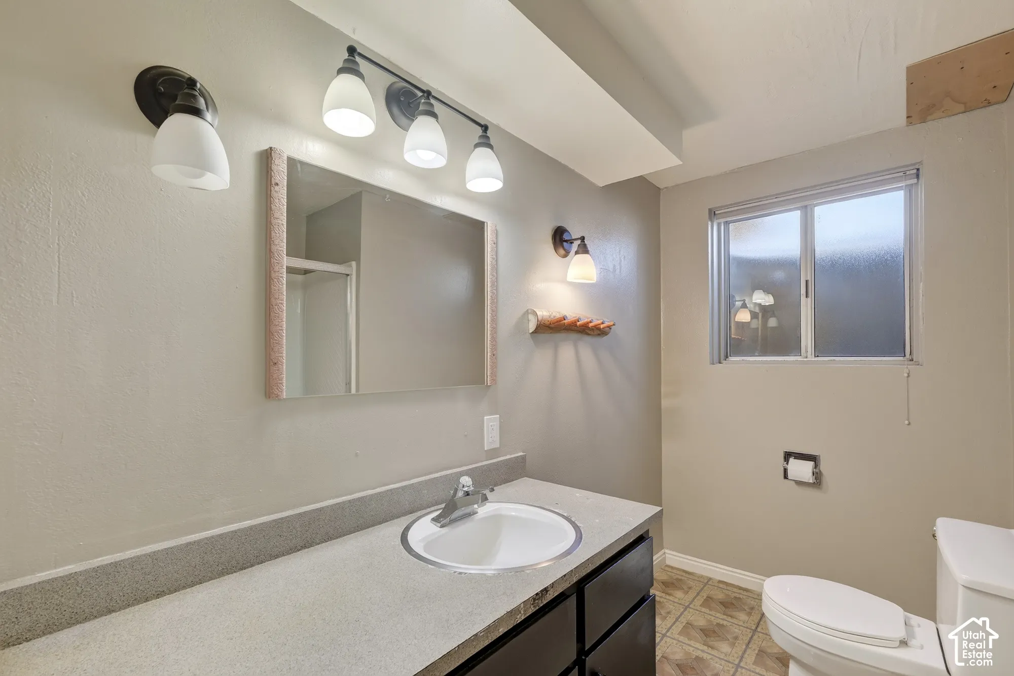 Bathroom vanity with stone-composite countertop, integrated sink, dark cabinetry, multi-bulb wall lighting, and obscured-glass window