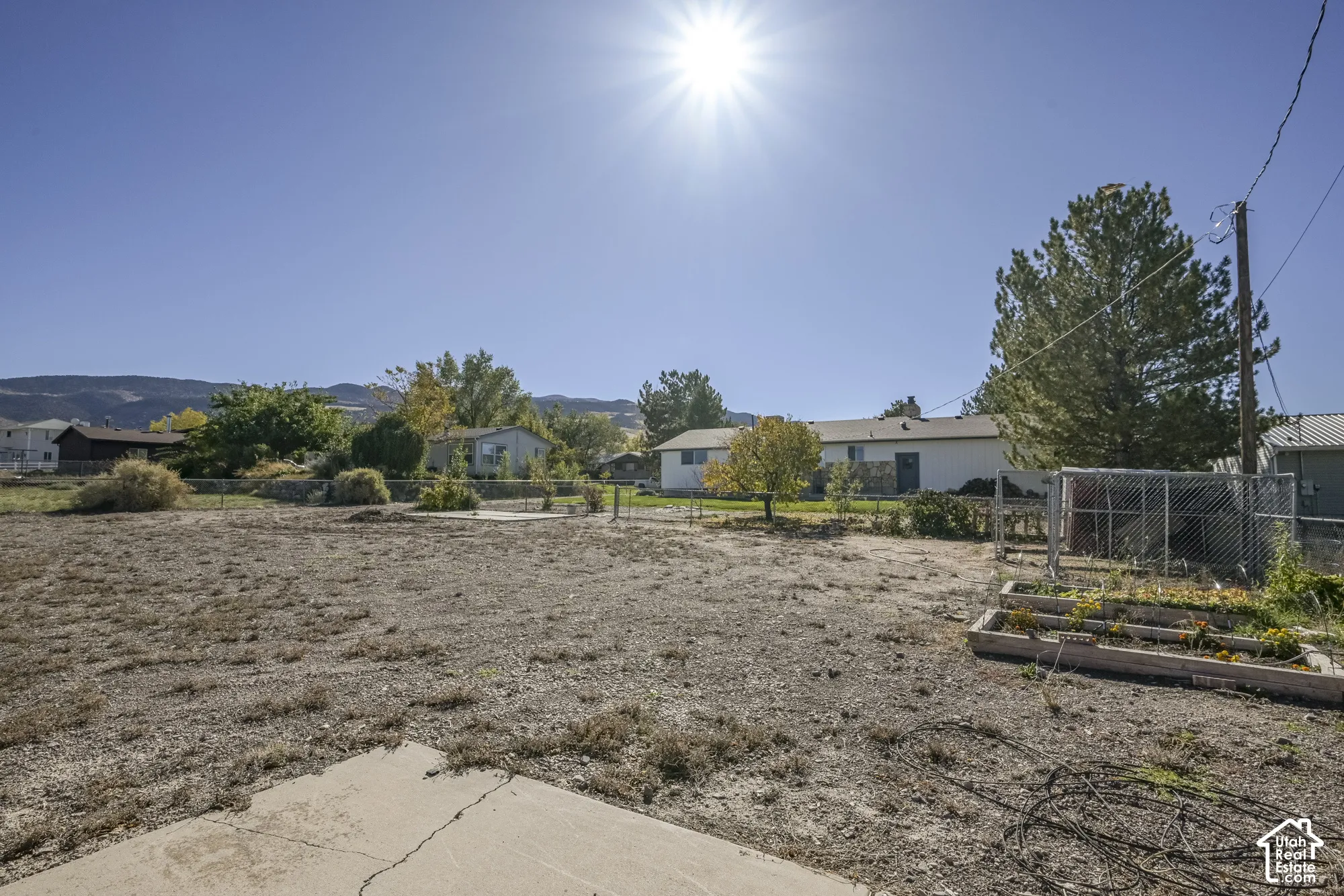 Expansive lot featuring a concrete patio, raised garden beds, chain-link fencing, and mature trees against a mountain backdrop