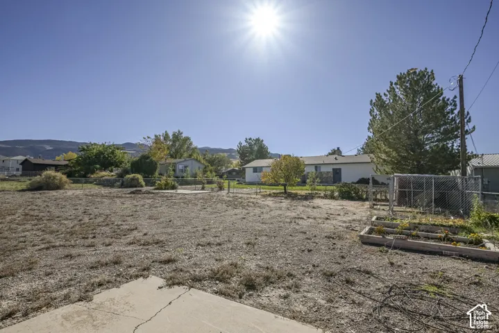 Expansive lot featuring a concrete patio, raised garden beds, chain-link fencing, and mature trees against a mountain backdrop