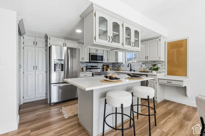 Kitchen featuring white cabinetry with glass-front uppers, an island with stone-finish countertops, stainless steel appliances, multi-tonal backsplash tile, and wood-finish flooring