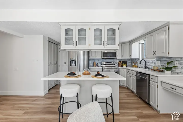 Kitchen featuring white shaker-style cabinetry with glass-front uppers, stainless steel appliances, a central island with a quartz countertop, multi-tonal tile backsplash, and wood-finish flooring