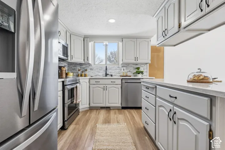 Kitchen featuring light-toned cabinetry, stainless steel appliances, stone-look tile backsplash, quartz countertops, and wood-finish flooring