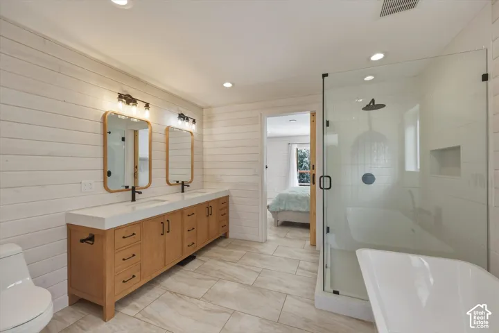 Dual vanity with integrated sinks, wood-finish cabinetry, floor-to-ceiling horizontal wall paneling, glass-enclosed walk-in shower with rain showerhead, and a standalone soaking tub