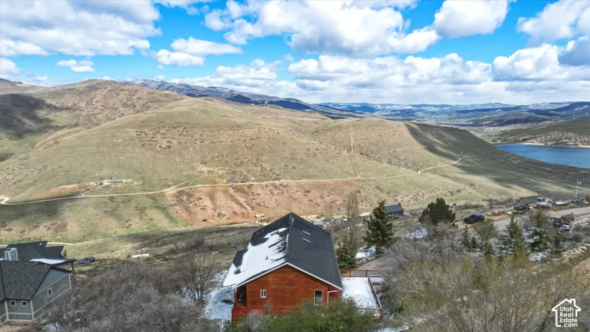 Aerial perspective of a multi-level wood-sided residence featuring a dark shingle roof with skylights, situated on an elevated lot with sweeping mountainous terrain and nearby water access