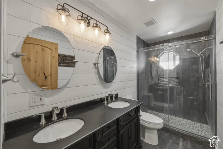 Double vanity with dark cabinetry, stone-finish countertop, dual circular mirrors, shiplap wall paneling, and a walk-in shower with glass enclosure and stone-tile flooring