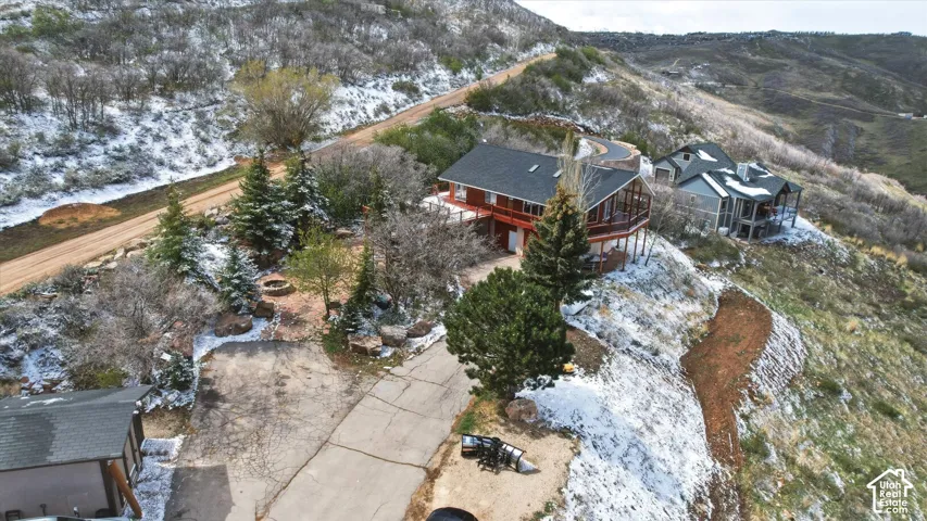 Elevated residence featuring a wrap-around deck, steep-gabled roofline, concrete driveway, stone-lined landscaping, and mature evergreen trees