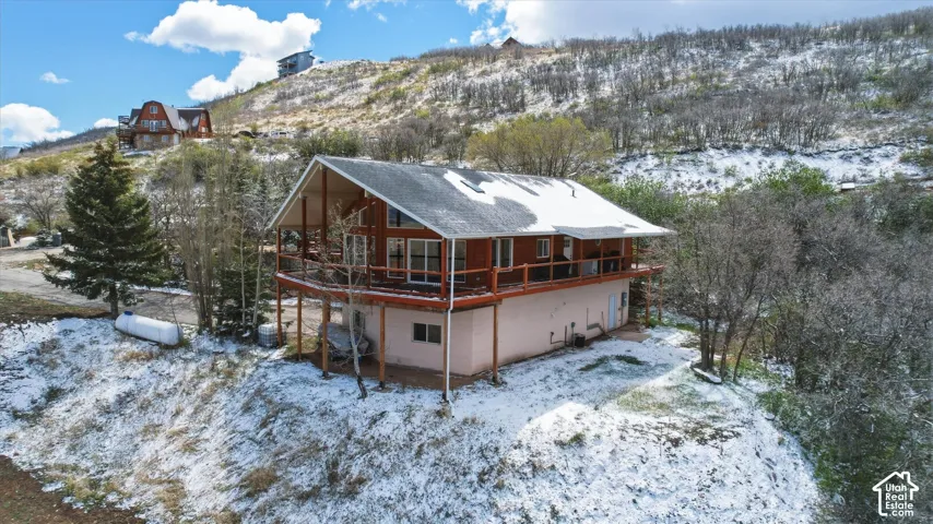 Two-story residence featuring wood-clad siding, a wraparound elevated deck, and a pitched roof, situated on a hillside with native trees and rugged terrain