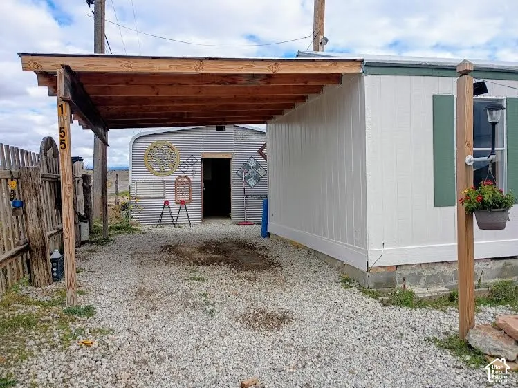 Covered carport featuring a natural wood beam frame and gravel surface, paired with a metal-sided exterior structure and wooden perimeter fencing