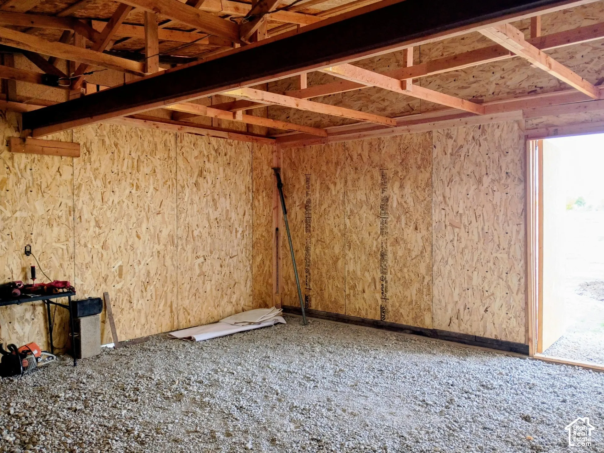 Interior space featuring exposed wood-truss ceiling, oriented strand board wall paneling, structural steel beam, and gravel flooring