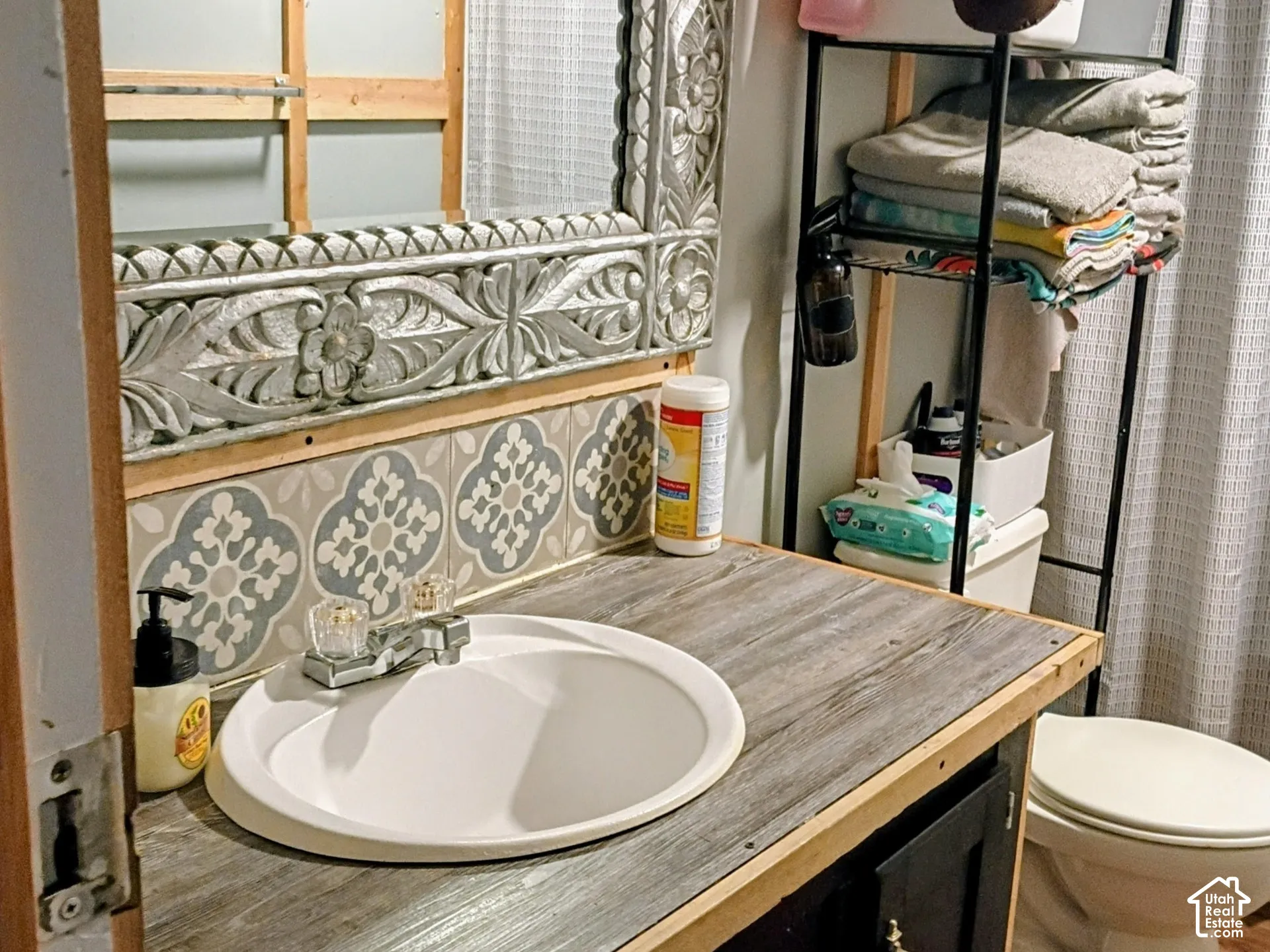 Bathroom vanity featuring a wood-finish countertop, white drop-in basin, decorative patterned tile backsplash, and dark cabinetry