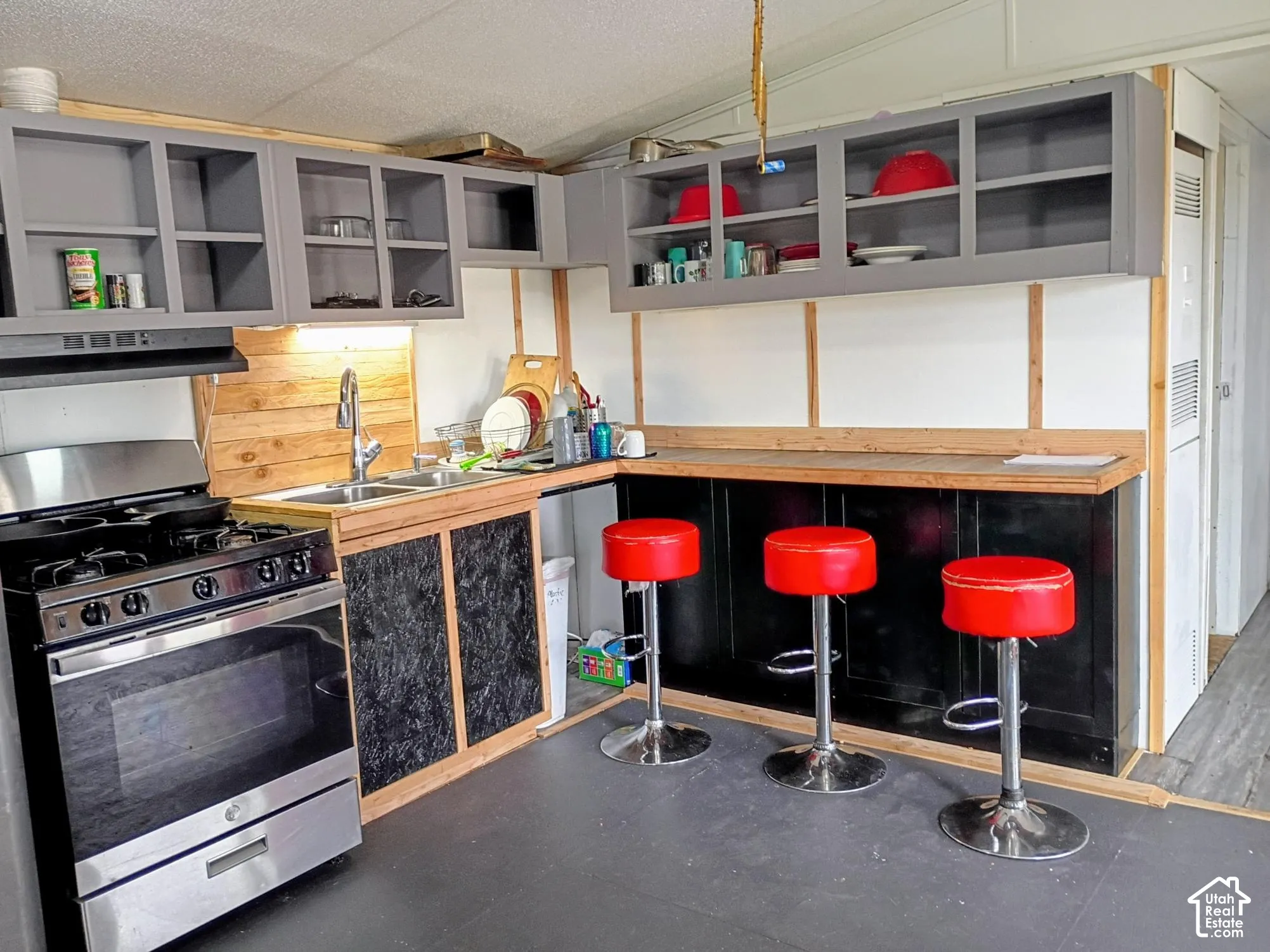 Kitchen featuring grey wall-mounted cabinetry, stainless steel gas range, wood-toned backsplash, double-basin sink with a gooseneck faucet, and an integrated counter-height breakfast bar
