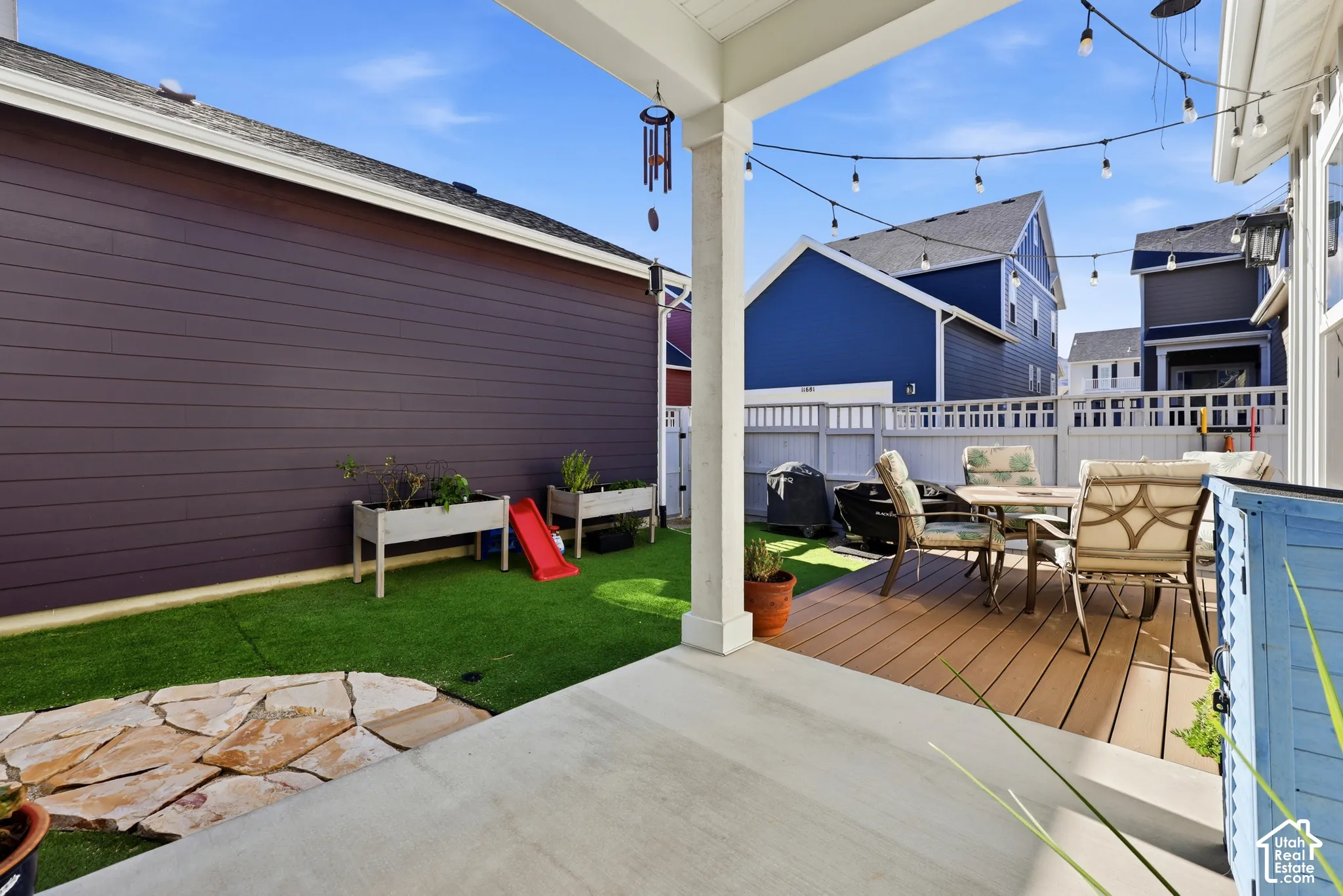 Covered concrete patio featuring synthetic turf, decorative flagstone hardscaping, and low-maintenance composite decking, all enclosed by white vinyl fencing