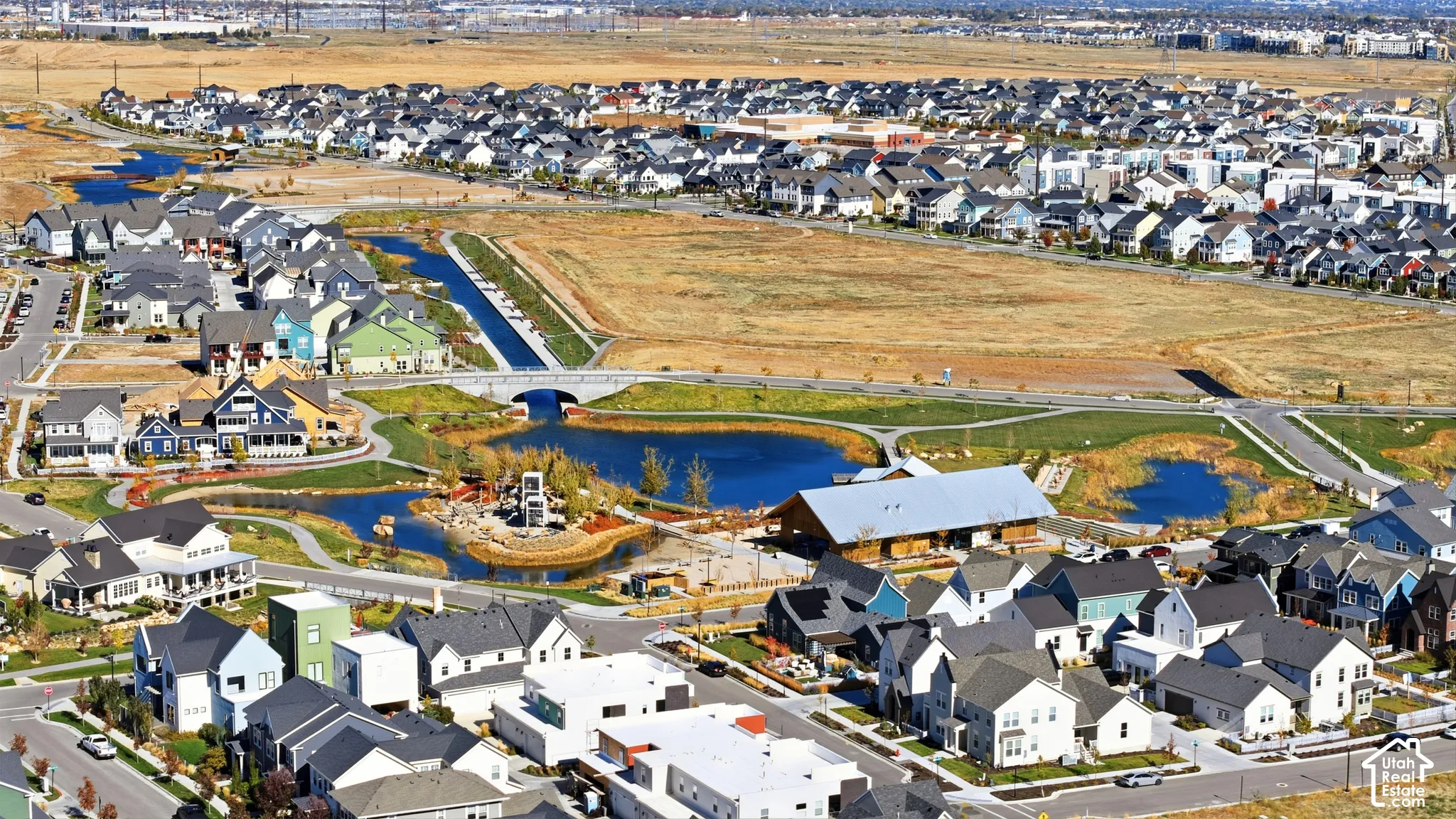 Aerial perspective of a master-planned community featuring multi-level residential architecture, central water features, pedestrian pathways, and a large-scale community building with a metal roof