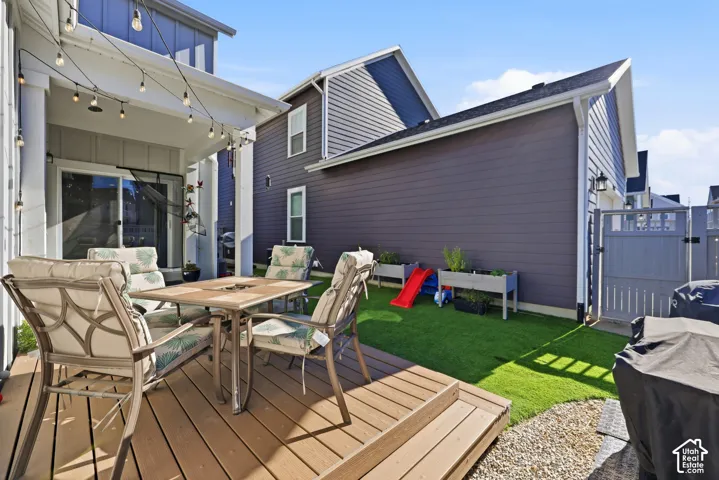 Covered patio featuring a wood-tone deck, synthetic turf lawn, horizontal lap siding, and a gated side access point