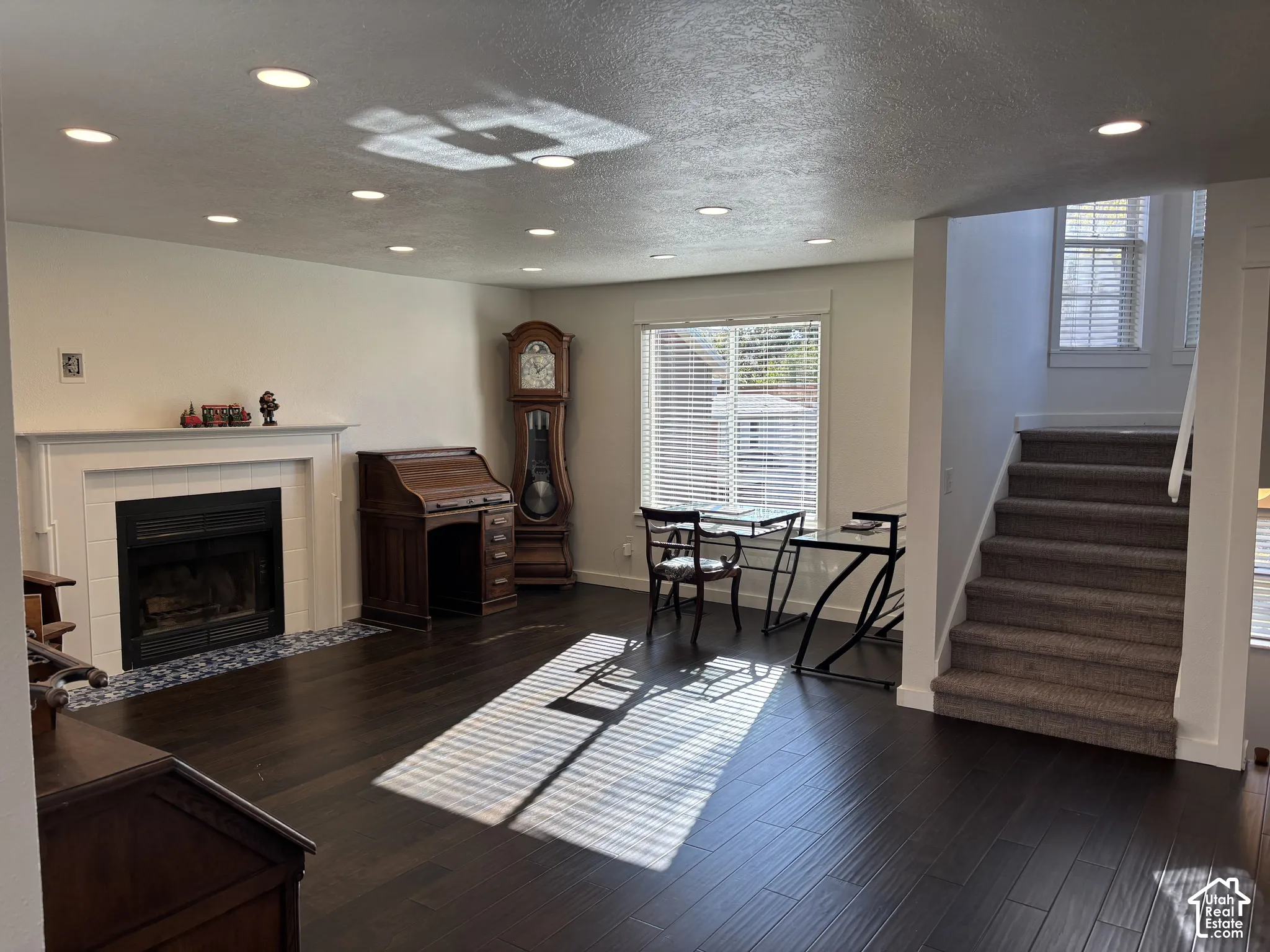 Dark wood-finish flooring, recessed lighting, fireplace with a white mantel and tile surround, and carpeted staircase