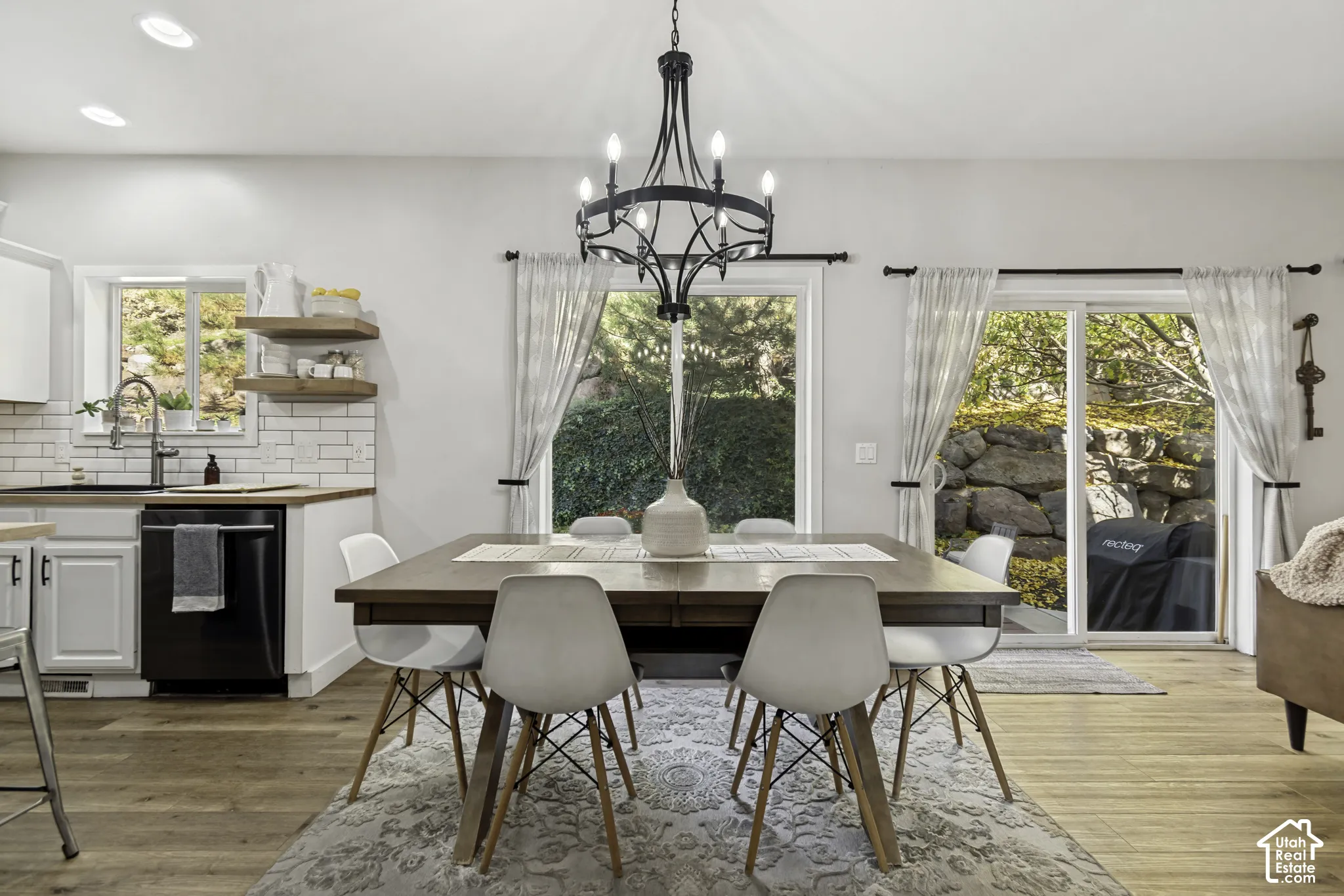 Open-concept space featuring wood-finish flooring, white subway tile backsplash, floating wood shelving, matte black chandelier, and sliding glass door