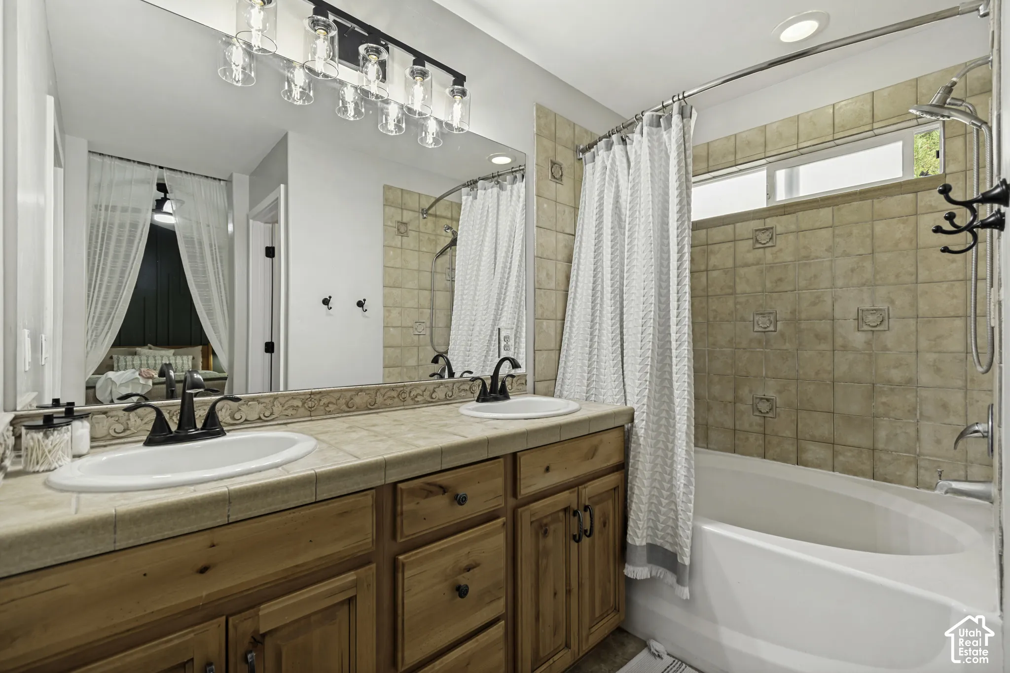 Dual-sink vanity with tile countertop, wood cabinetry, large vanity mirror, multi-light fixture, and tile-surround soaking tub