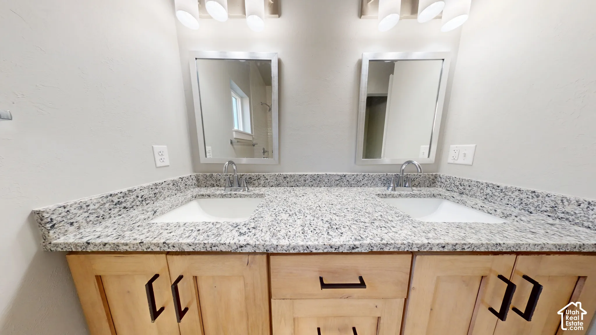 Dual-sink vanity featuring light-toned wooden cabinetry, stone-surface countertop, rectangular undermount basins, and dark metal hardware