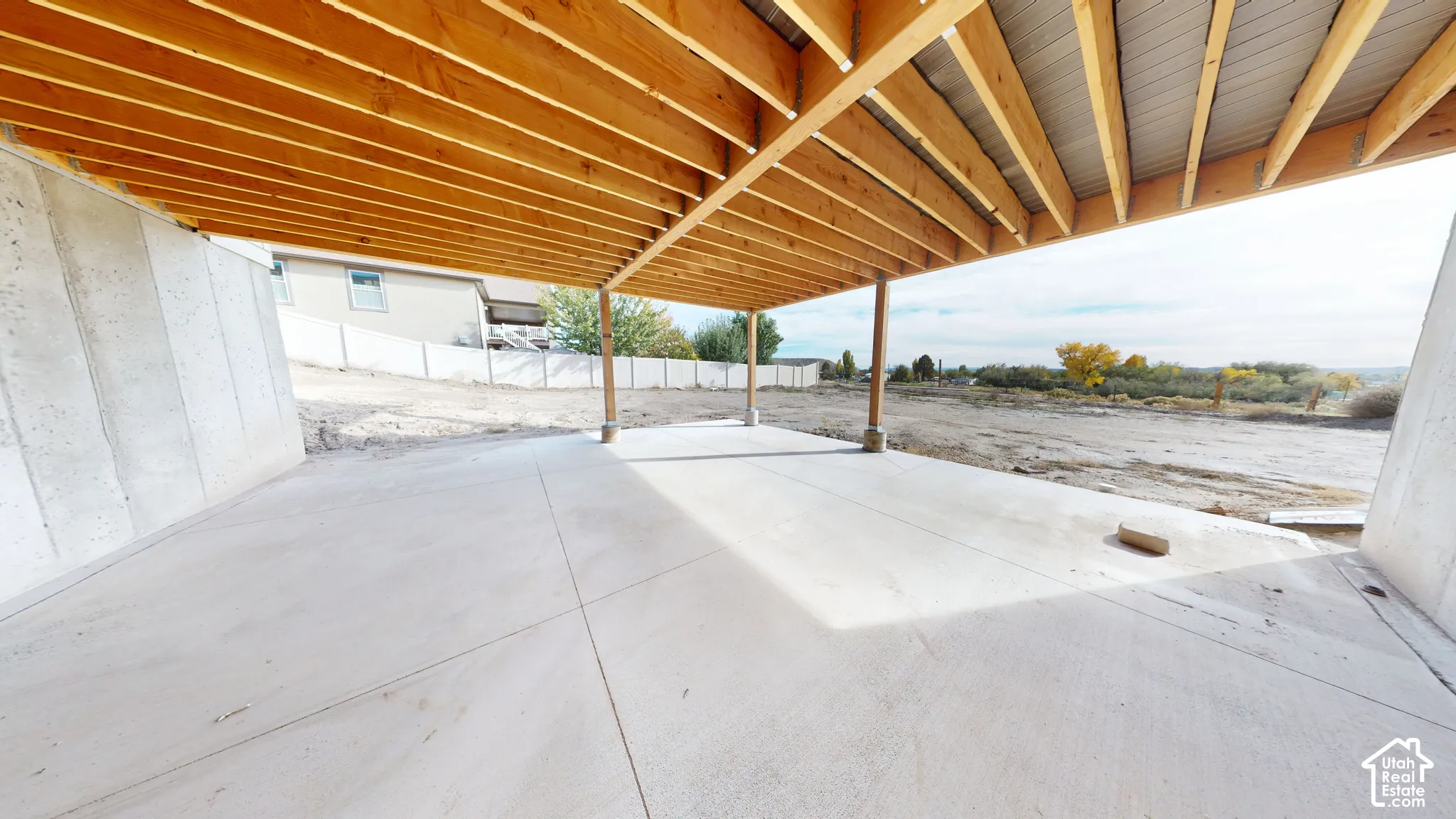 Covered concrete patio featuring exposed natural wood-finish ceiling joists and support beams, cast-in-place concrete foundation walls, and level ground area