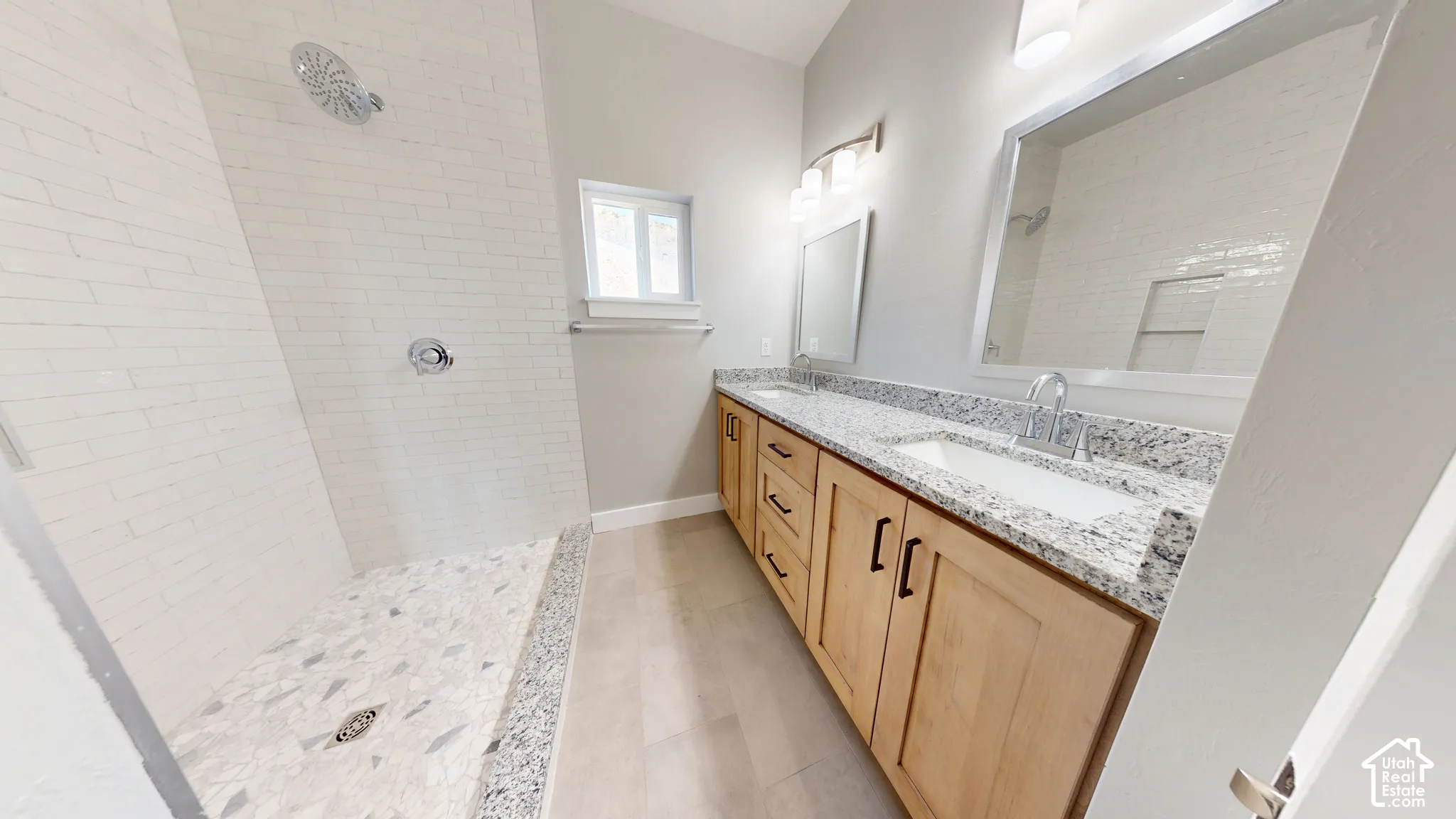 Bathroom featuring a dual-sink vanity with light wood cabinetry, stone-surface countertops, a walk-in shower with a mosaic tile floor, and white subway tile wall surrounds