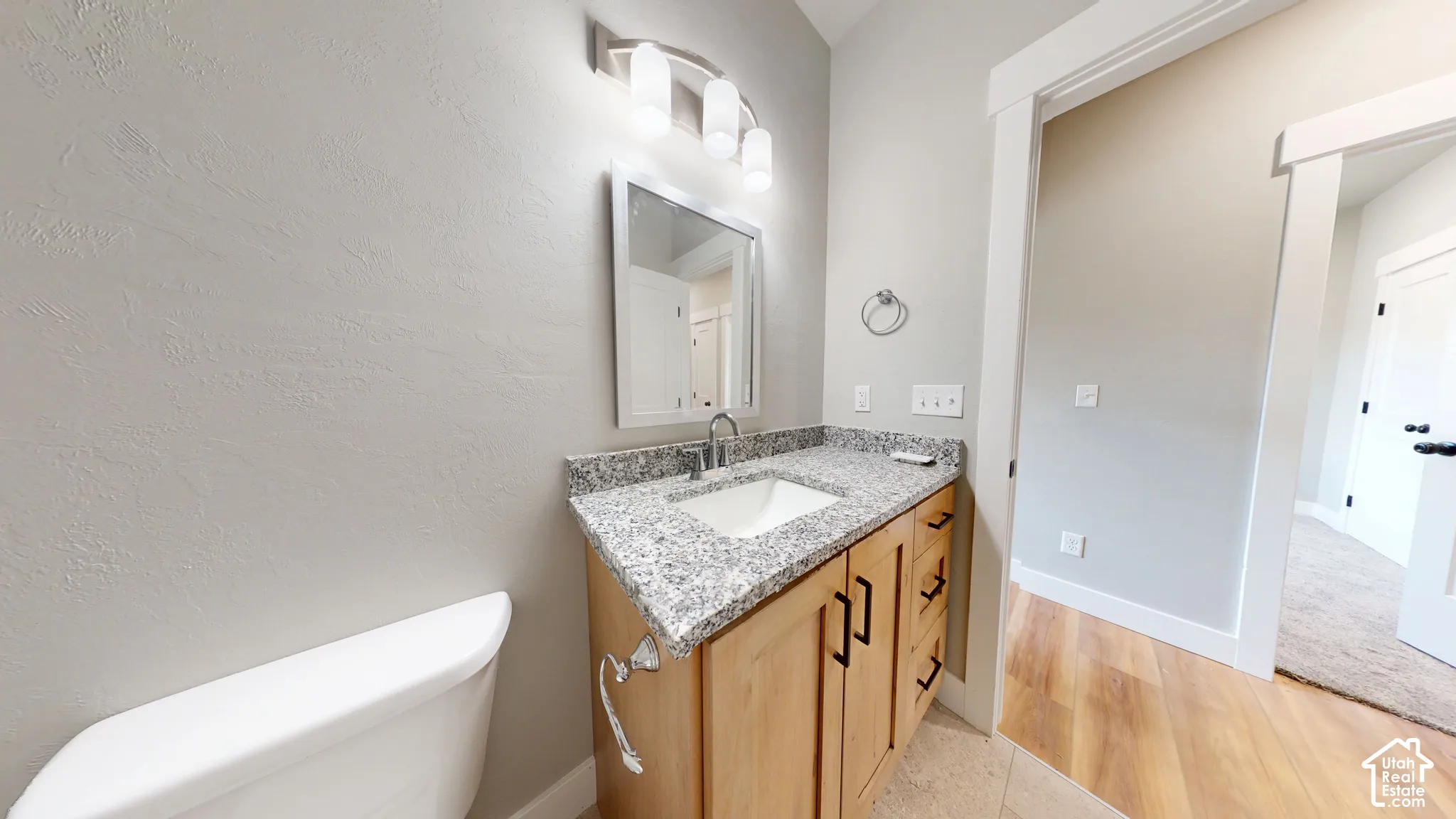 Vanity featuring light wood-finish cabinetry, stone-composite countertop with an integrated basin, brushed metal hardware, and a three-light wall-mounted fixture