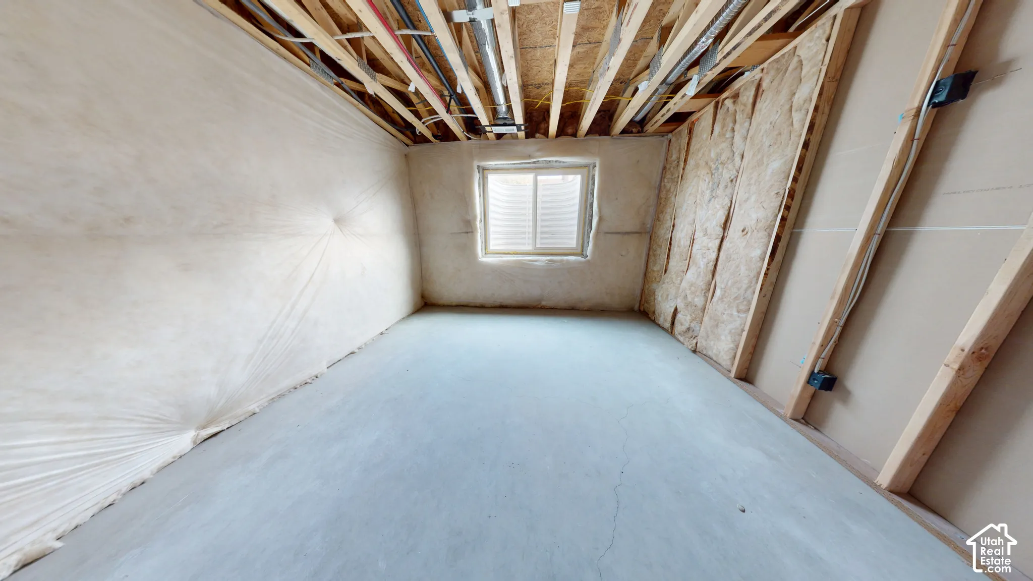 Unfinished room featuring exposed wooden ceiling joists, concrete flooring, insulation-wrapped walls, and a recessed window