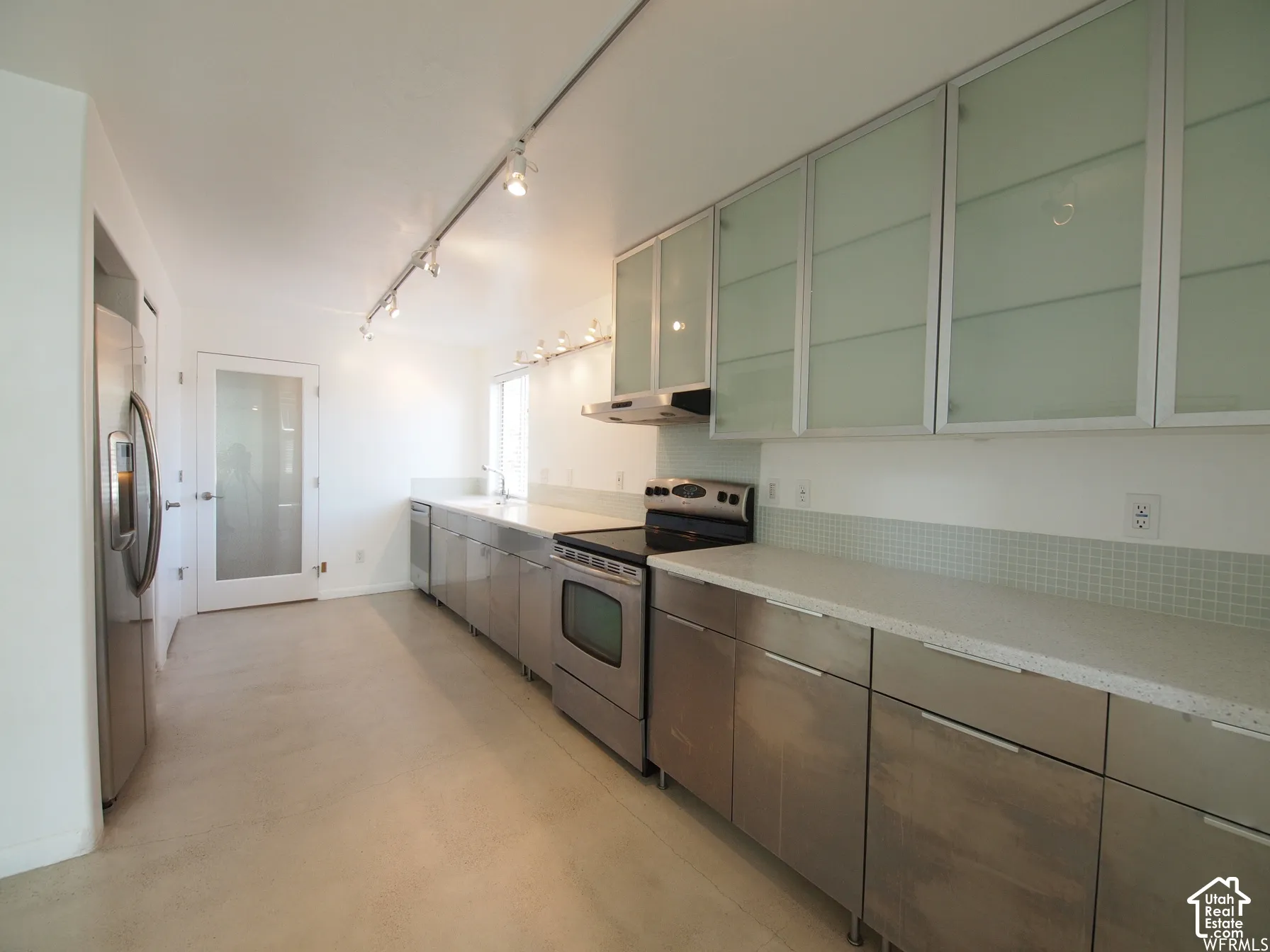 Kitchen featuring stainless steel cabinetry, light-toned stone countertops, frosted glass upper cabinet doors, mosaic tile backsplash, and track lighting