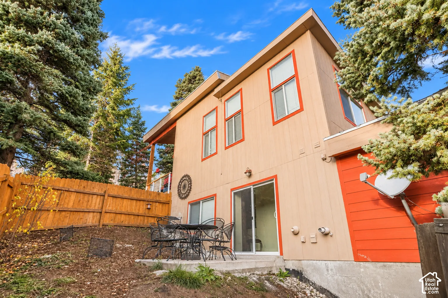 Two-story exterior featuring vertical wood siding, concrete patio, sliding glass door, and wooden perimeter fencing surrounded by mature evergreen trees