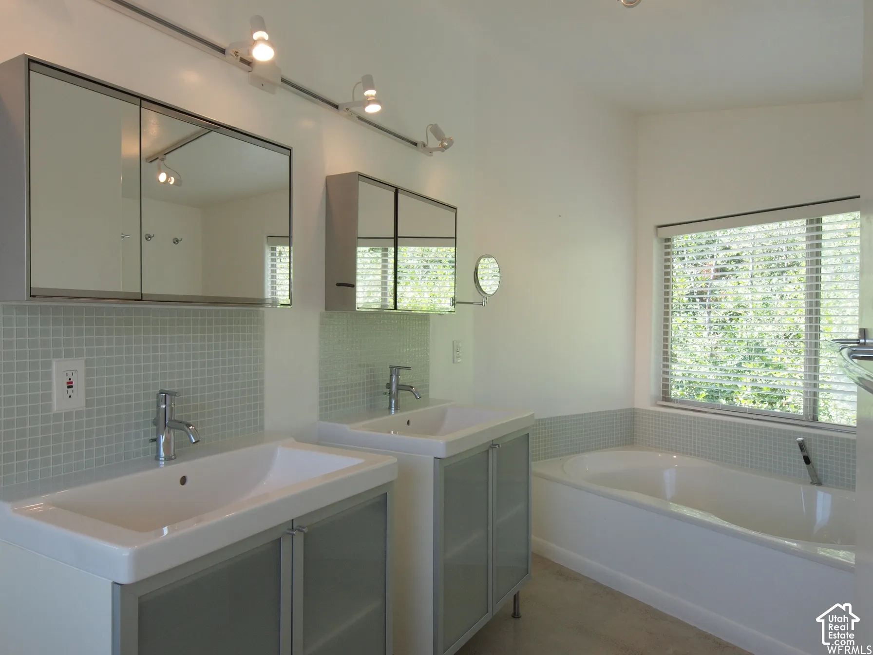 Dual vanity featuring glass-front cabinetry, integrated rectangular sinks, polished chrome faucets, wall-mounted mirrored medicine cabinets, and a deep soaking tub with a tiled surround