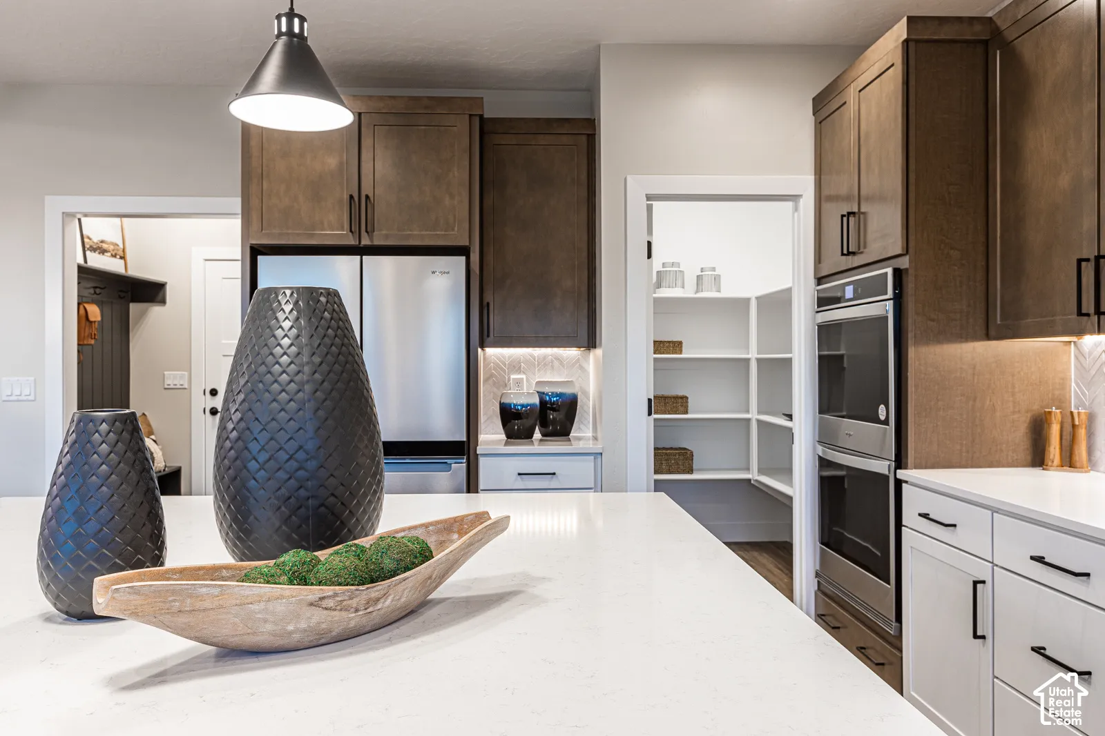 Kitchen featuring white solid-surface countertops, custom stained cabinetry, built-in double wall ovens, stainless steel refrigerator, and a walk-in pantry with built-in shelving