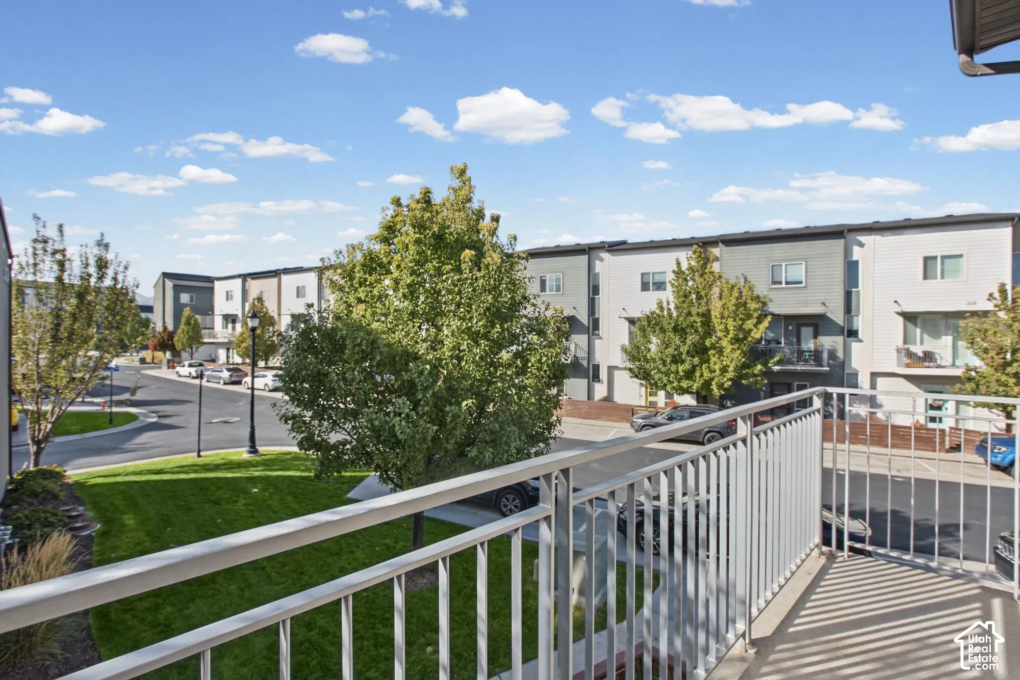 Elevated balcony with a metal railing overlooking a community streetscape, featuring mature trees and landscaped green space