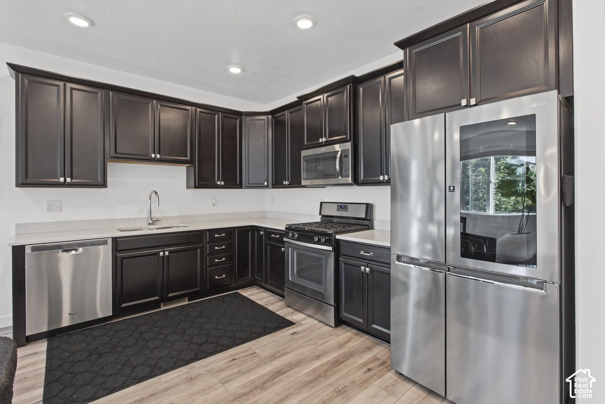 Kitchen featuring espresso-toned cabinetry, stainless steel appliances, light-toned stone-surface countertops, wood-finish flooring, and recessed ceiling lighting