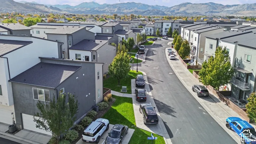 Aerial perspective of a multi-unit residential community featuring asphalt-paved streets, concrete sidewalks, mature deciduous trees, and a mountain range backdrop