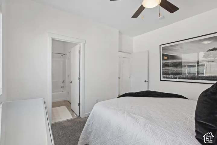 Neutral-toned interior featuring carpeted flooring, a ceiling fan with integrated lighting, and a white interior door opening to an en suite featuring a tub and shower surround