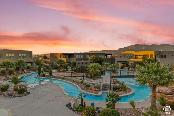 Community aquatic feature with winding lazy river, concrete decking, metal pedestrian bridges, mature palm trees, and desert landscape accents set against a mountain backdrop