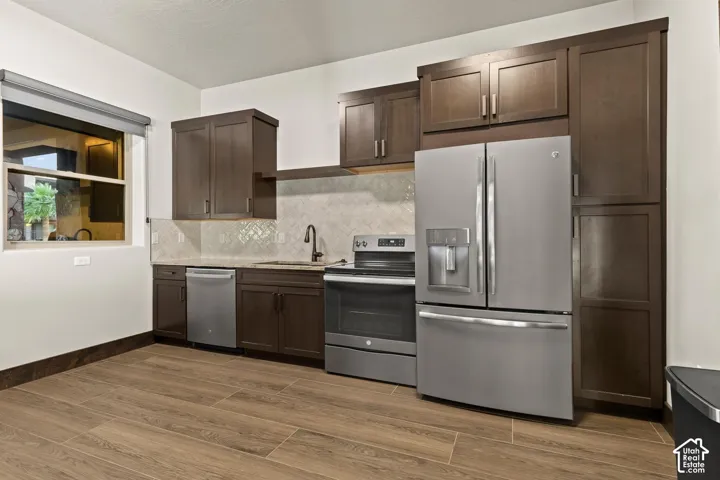 Kitchen featuring dark wood cabinetry, stainless steel appliances, neutral stone-finish countertops, herringbone-pattern tile backsplash, and wood-finish flooring