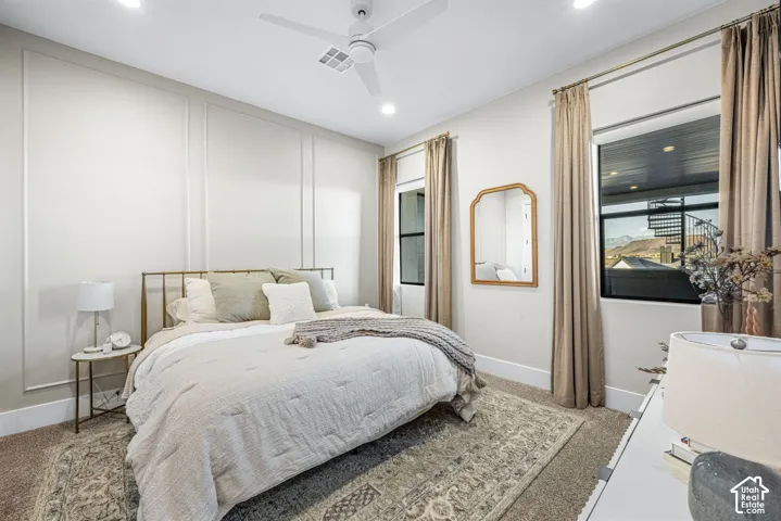 Bedroom featuring decorative wall molding, a modern white ceiling fan with integrated lighting, dual windows with black frames, and neutral carpeting