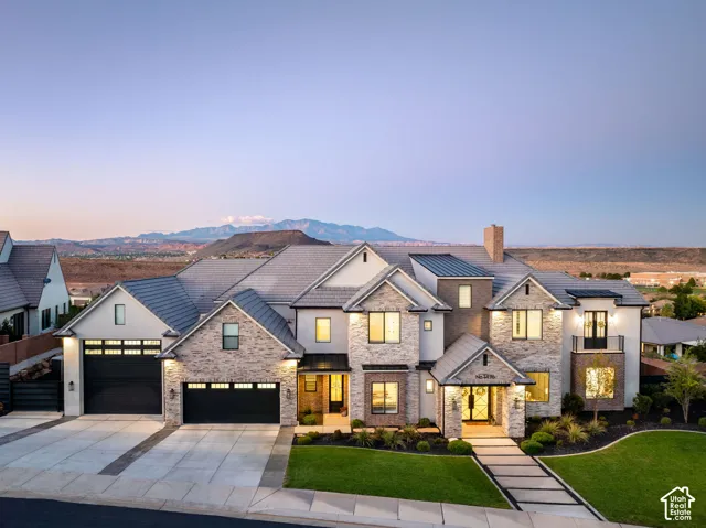 Multi-gabled exterior featuring stone masonry and stucco siding, tile roof, expansive multi-car garage with glass-paneled doors, paved walkway with integrated landscaping, and mountain backdrop