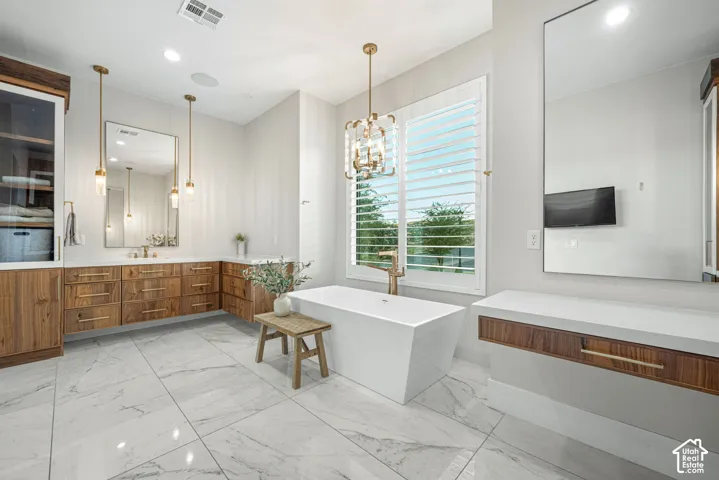Freestanding rectangular soaking tub with a wall-mounted faucet, custom wood-finish cabinetry, quartz countertops, large-format tile flooring, and decorative pendant lighting